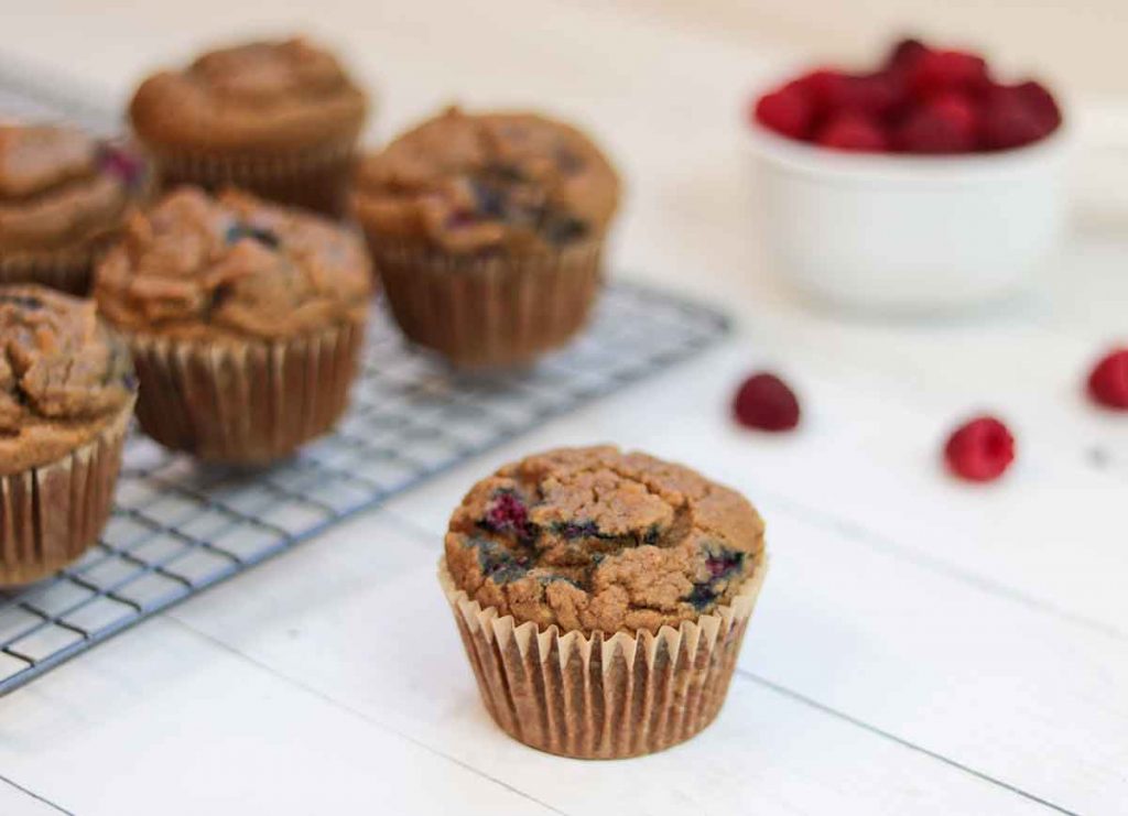 1 raspberry muffin with the rest of the muffins and a bowl of raspberries behind