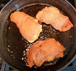 three pieces of chicken frying in cast iron pan