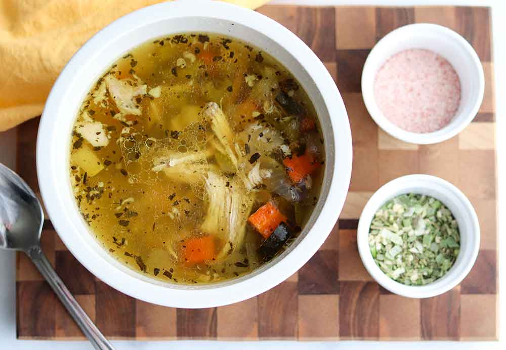 Crockpot chicken soup in a white ceramic container placed on a cutting board beside spices