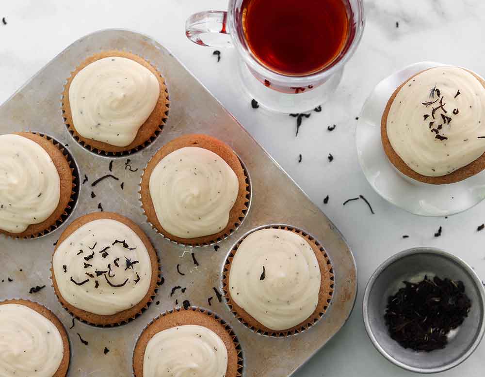 Early grey cupcakes sitting in muffin tin topped with icing and placed beside earl grey tea and a plated muffin