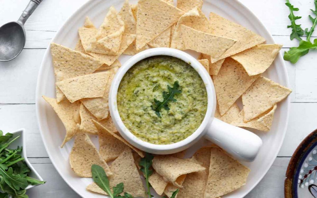 Green artichoke dip placed on a white plate covered in cassava flour chips and surrounded by fresh greenery and clean slat boards