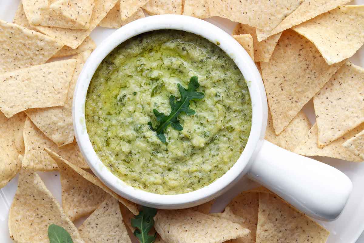 Close up view of green artichoke dip in a white ceramic dish surrounded by cassava flour chips