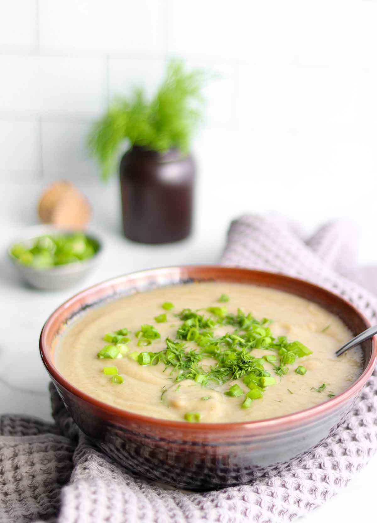 Creamy cauliflower soup in a brown bowl with fresh herbs placed in backdrop