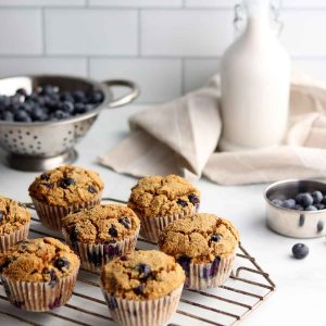 side view of tiger nut blueberry muffins placed on a wire cooling rack with a colander of blueberries and milk alternative in the background