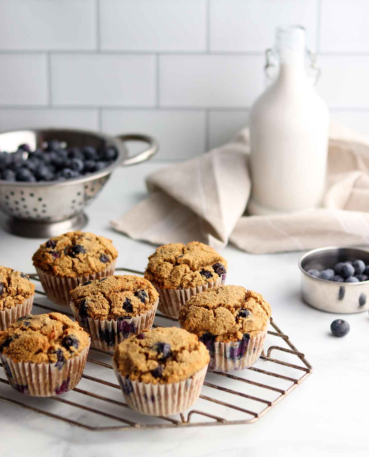 side view of tiger nut blueberry muffins placed on a wire cooling rack with a colander of blueberries and milk alternative in the background
