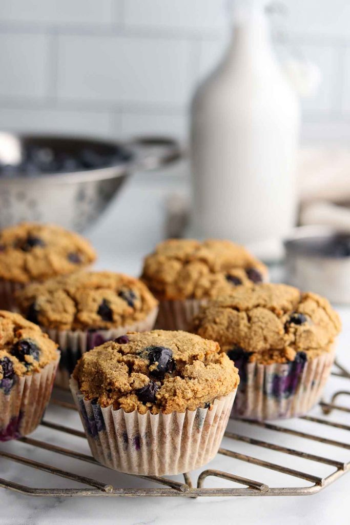 close up view of tiger nut blueberry muffins on wire cooling rack with a colander of blueberries in the back ground and a glass jar filled with a milk alternative