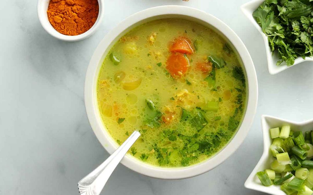 Close up overhead view of turmeric chicken soup in a white porcelain bowl surrounded by ingredients placed in decorative bowls