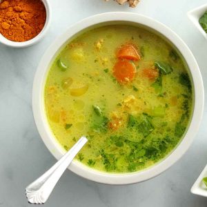 Close up overhead view of turmeric chicken soup in a white porcelain bowl surrounded by ingredients placed in decorative bowls