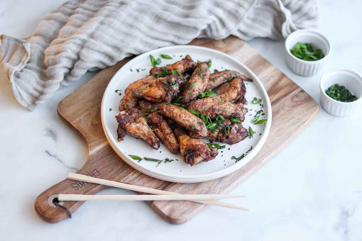 Close up of five spice chicken wings on a white ceramic plate ontop of a wooden cutting board sprinkled with green onion sitting beside a pair of chopsticks 