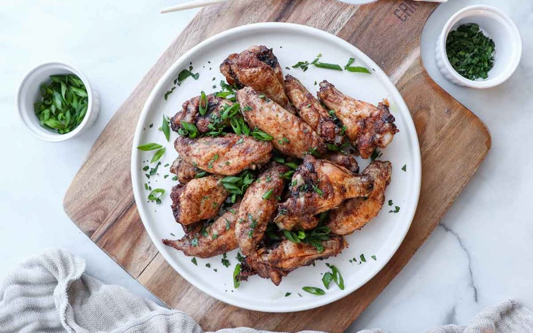 Overhead view of five spice chicken wings on a white ceramic plate placed ontop of a wooden cutting board surrounded by fresh and colorful ingredients