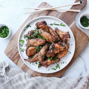 Overhead view of five spice chicken wings on a white ceramic plate placed ontop of a wooden cutting board surrounded by fresh and colorful ingredients