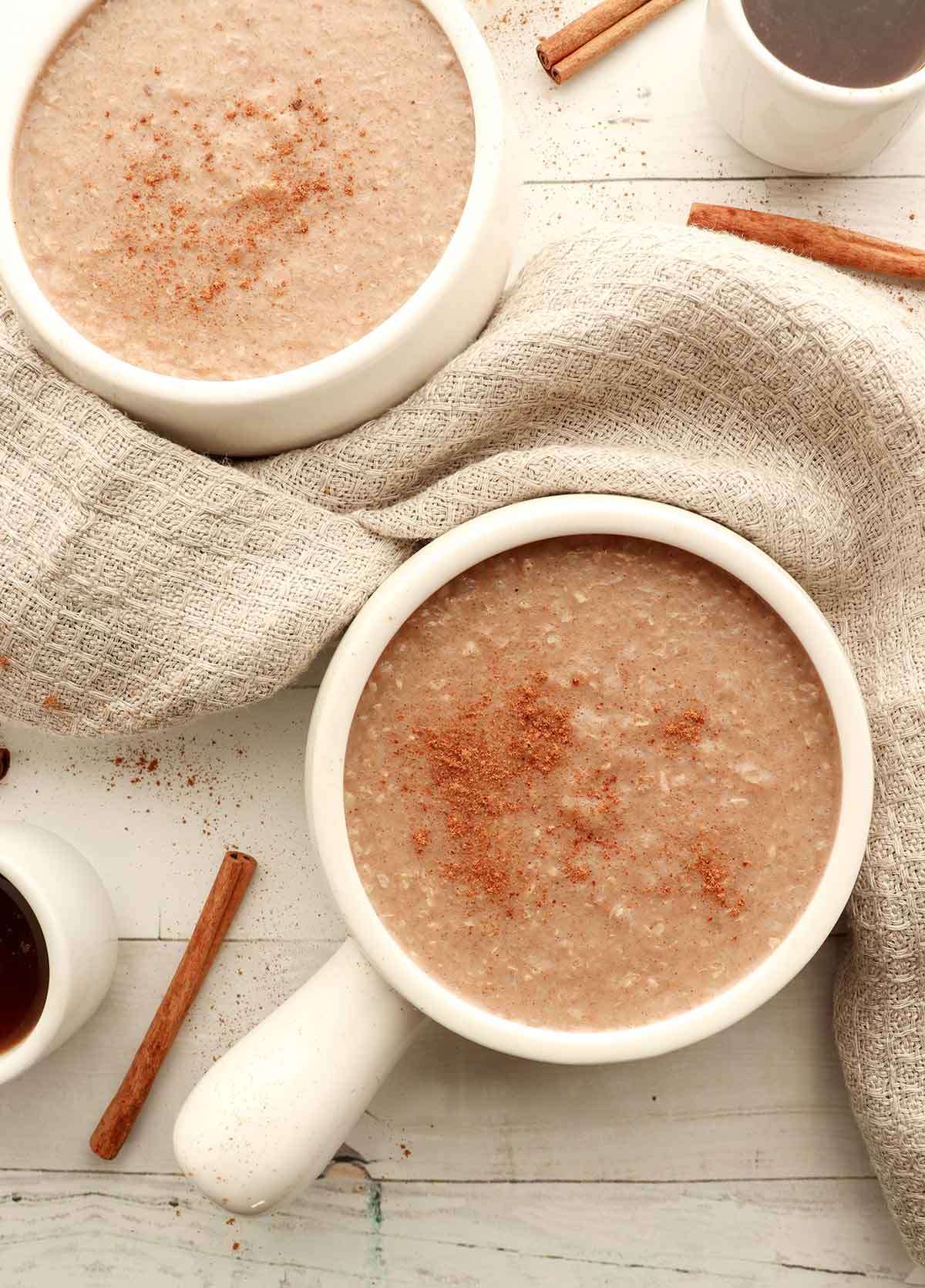 Overhead view of buckwheat porridge in two white ceramic dishes surrounded by cream coloured kitchen clothes, cinnamon sticks and coffee