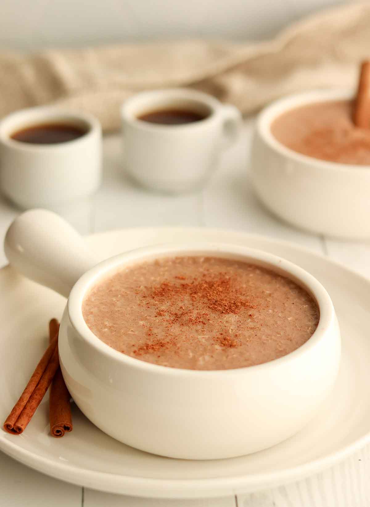 Close up view of buckwheat porridge in a white ceramic dish placed next to two coffee cups and a cinnamon stick 