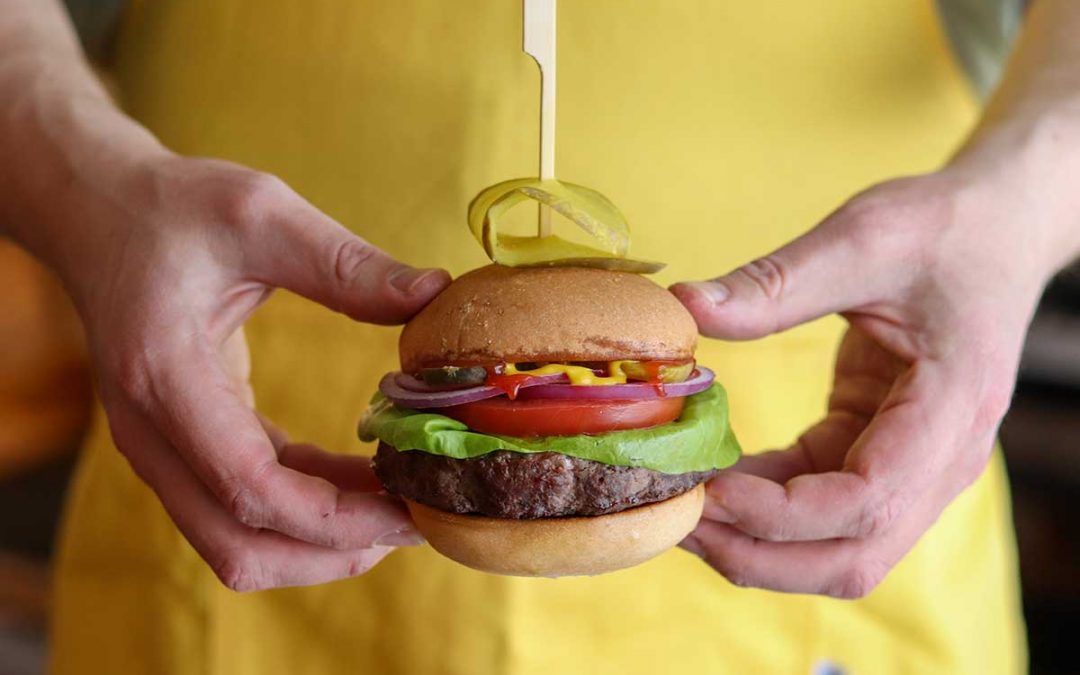Closed bun elk burger with all the toppings being held in front of a person wearing a yellow shirt