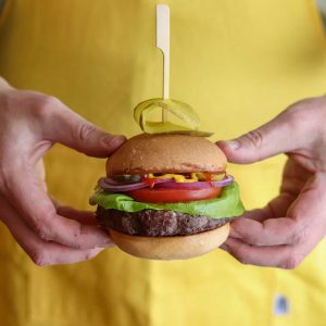 Closed bun elk burger with all the toppings being held in front of a person wearing a yellow shirt