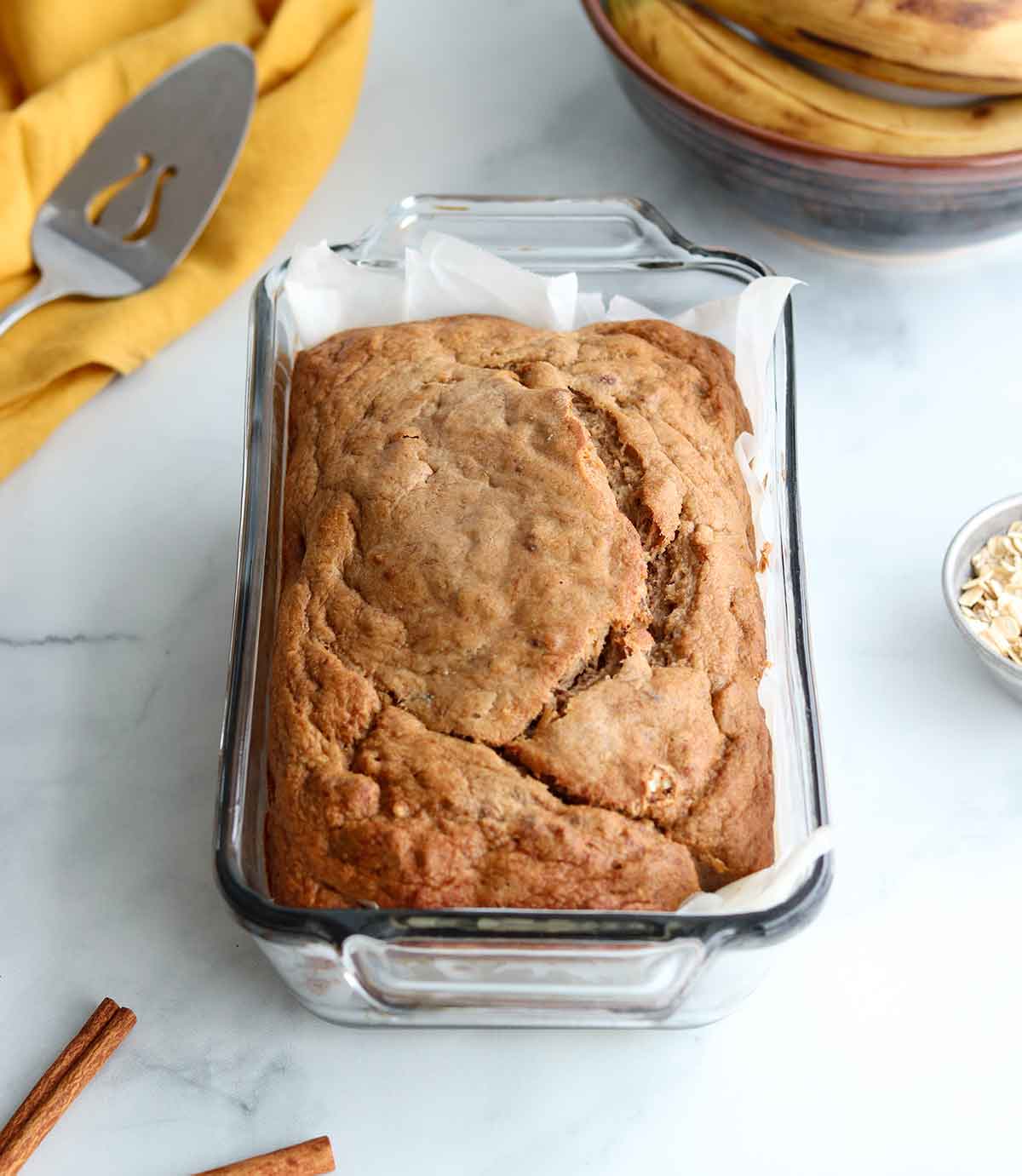 Oat flour banana bread in glass baking dish surrounded by baking necessities and ingredients 