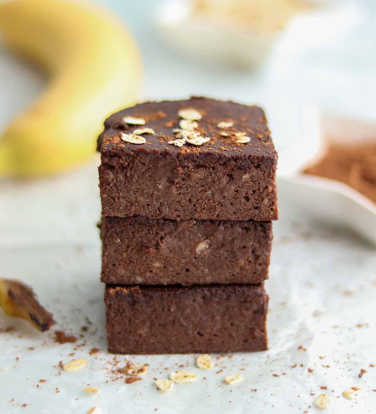 Oat flour brownie squares stacked on top of each other on oat dusted parchment paper, with a bowl of cocoa and a banana
out of focus in the background