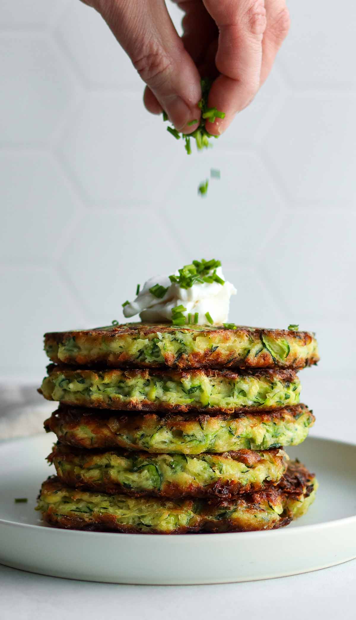 Close up view of a stack of zucchini fritters with a dollop of dairy free sour cream being sprinkled by green chives 