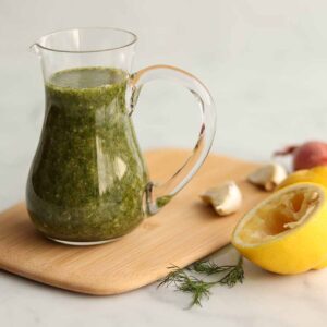 Close up view of lemon dill dressing in a glass pouring jar placed on top of a wooden serving board surrounded by fresh ingredients