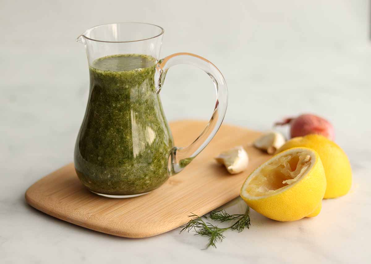 Close up view of lemon dill dressing in a glass pouring jar placed on top of a wooden serving board surrounded by fresh ingredients