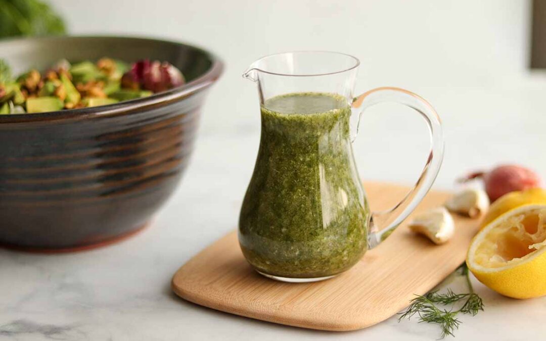 Lemon dill dressing placed on top of a wooden cutting board sitting beside a bowl of salad and garnishes