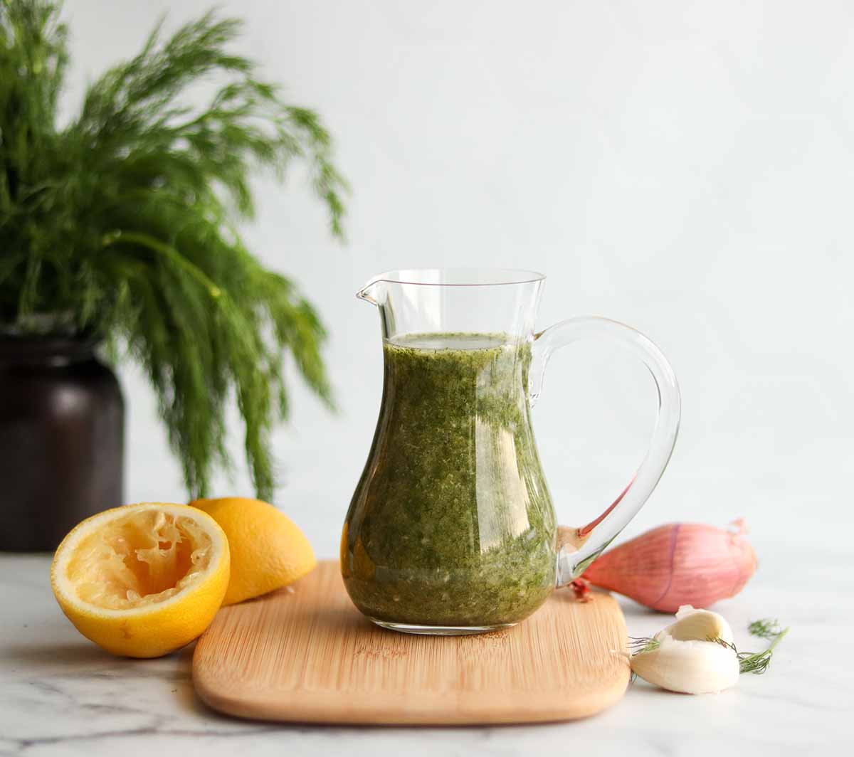 Close up view of lemon dill salad dressing in a glass pouring jar placed on top of a wooden cutting board surrounded by a large bunch of fresh dill and other ingredients