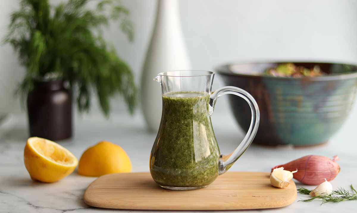 Panoramic side view of dill dressing in a glass pouring jar placed on top of a wooden serving board surrounded by fresh ingredients