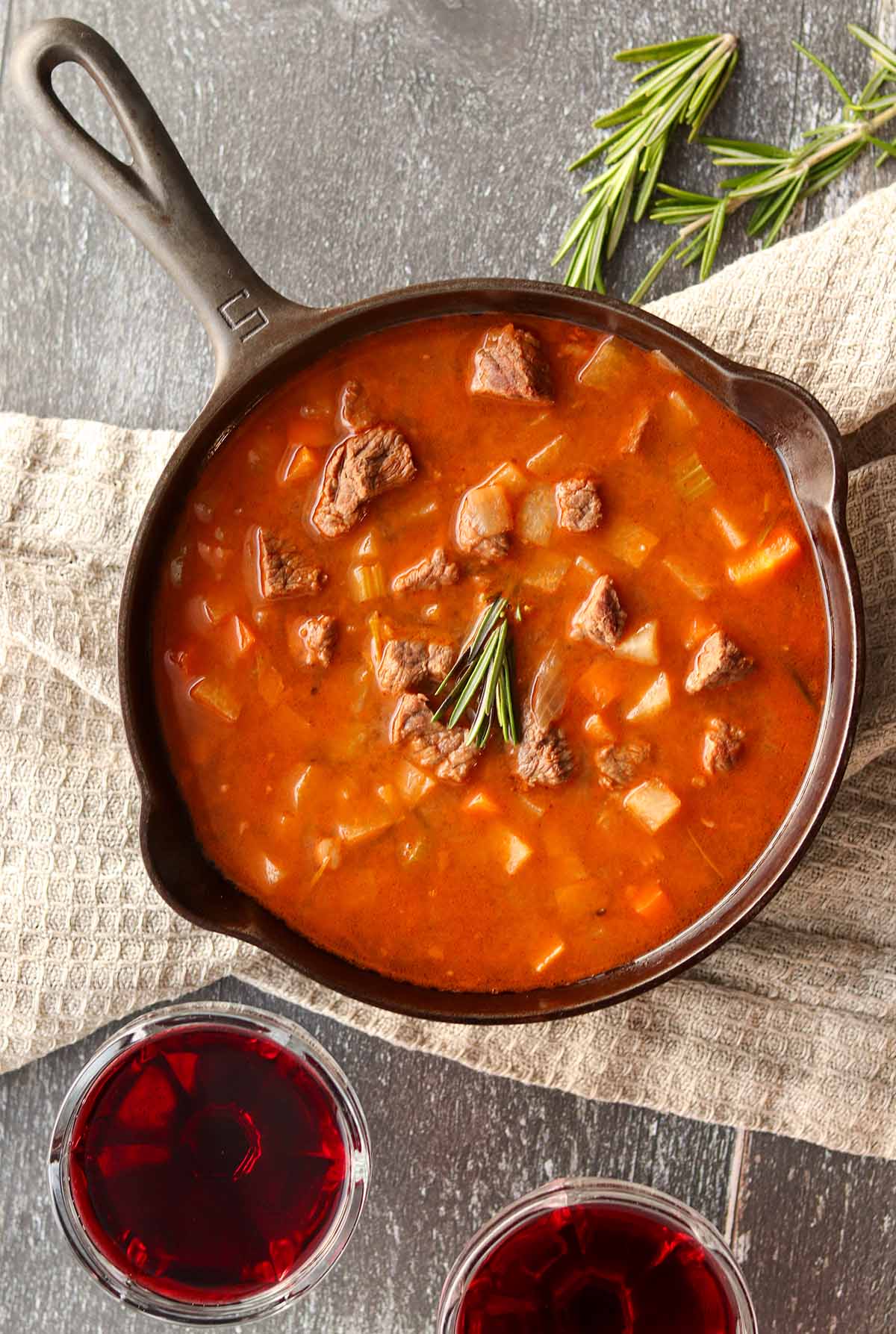 Overhead view of beef stew in a cast iron skillet surrounded by linen clothe, fresh herbs and two glasses of red wine