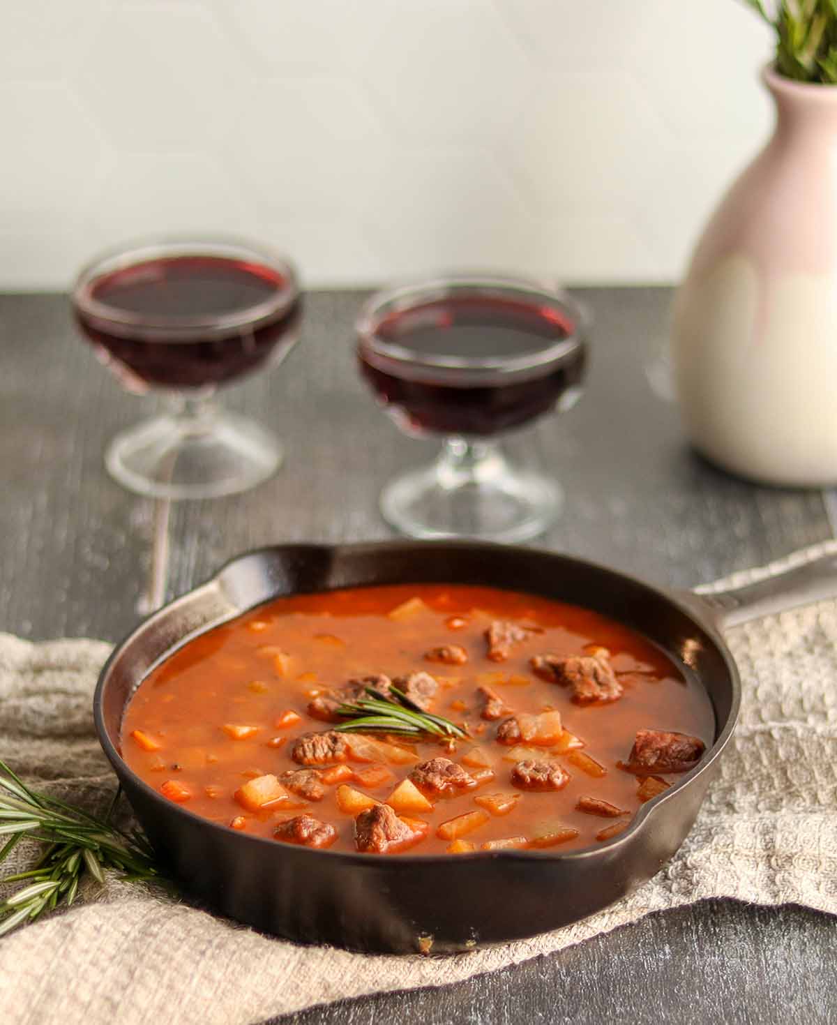 Side view of beef stew in cast iron skillet placed on top linen cloth and surrounded by fresh herbs and two glasses of wine