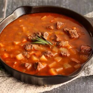 Side view of beef stew in cast iron skillet surrounded by herbs and linen clothe