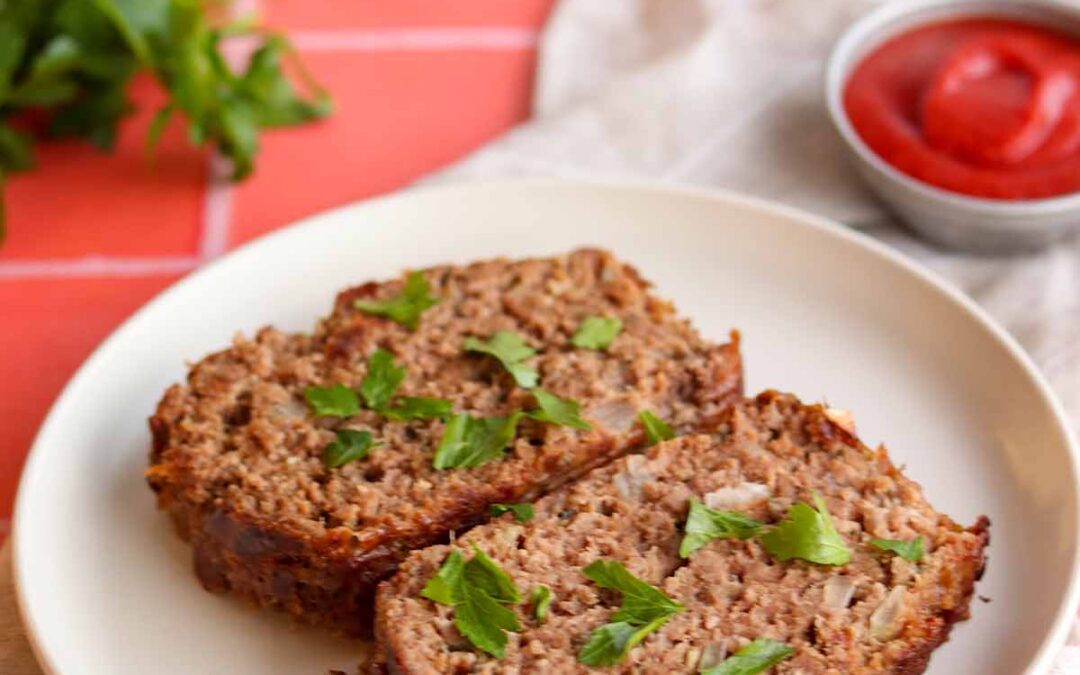 Overhead side view of paleo meatloaf, placed on top of a linen cloth draped across a red checkered table clothe and surround by ingredients