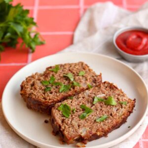 Overhead side view of paleo meatloaf, placed on top of a linen cloth draped across a red checkered table clothe and surround by ingredients