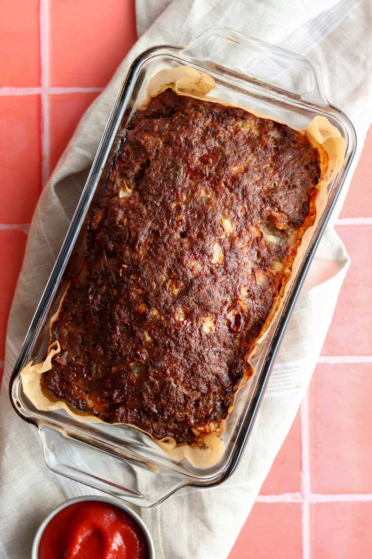 Overhead view of meat loaf in glass baking dish placed on top of a table cloth beside a ramekin of ketchup