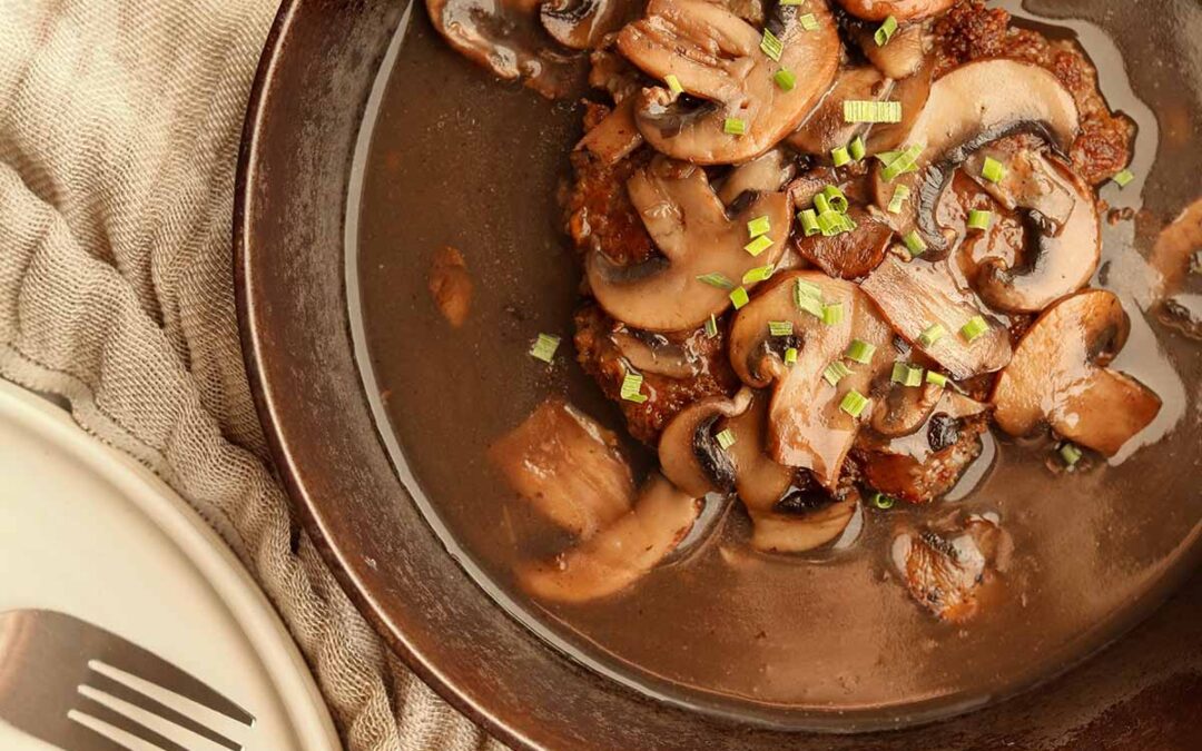 Close up view of salisbury steak in a cast iron skillet on top of a linen cloth beside a ceramic plate and fork