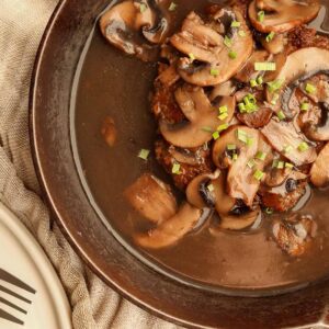 Close up view of salisbury steak in a cast iron skillet on top of a linen cloth beside a ceramic plate and fork