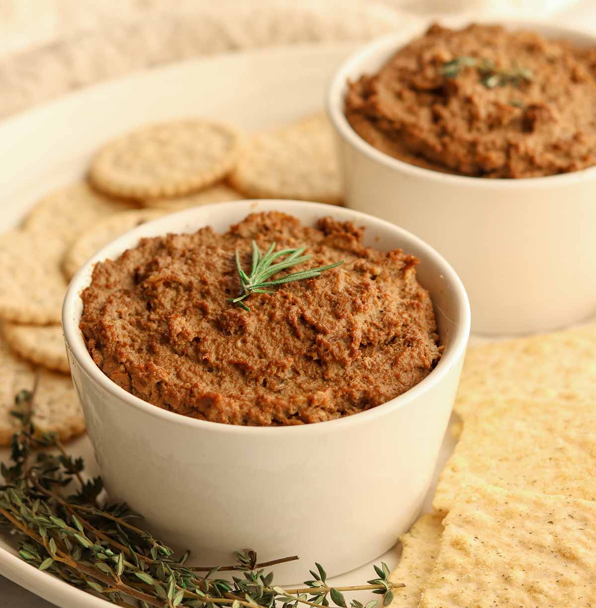 Side view of beef liver pate in two white ceramic dishes surrounded by crackers and garnish