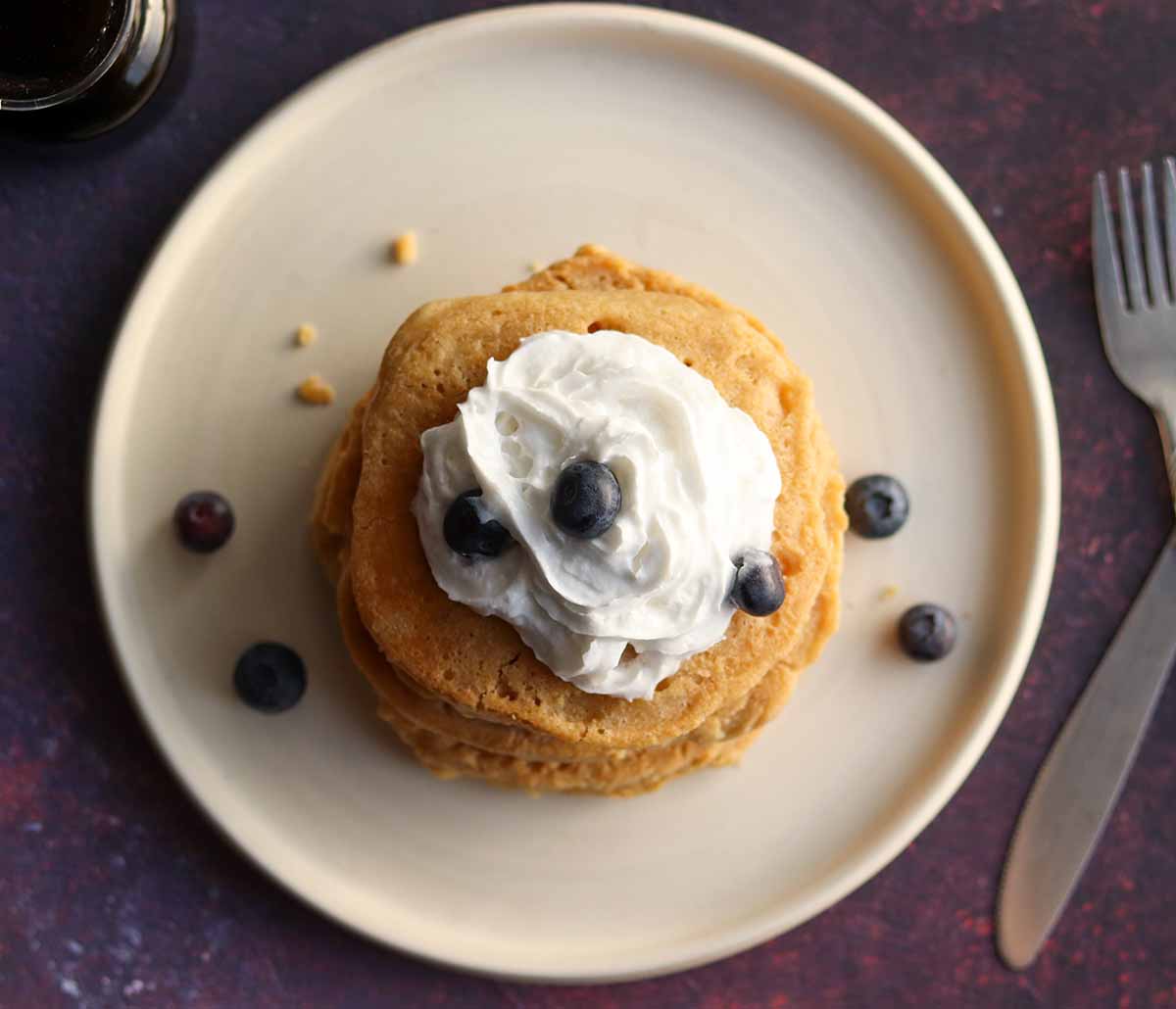 Overhead view of stacked oat flour pancakes with toppings on a ceramic plate surrounded by cutlery