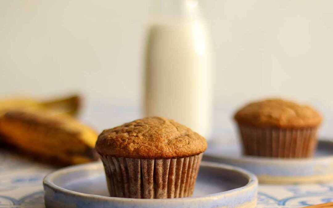 Centered close up of paleo banana muffin on blue ceramic plate surrounded by ingredients and other muffin in background