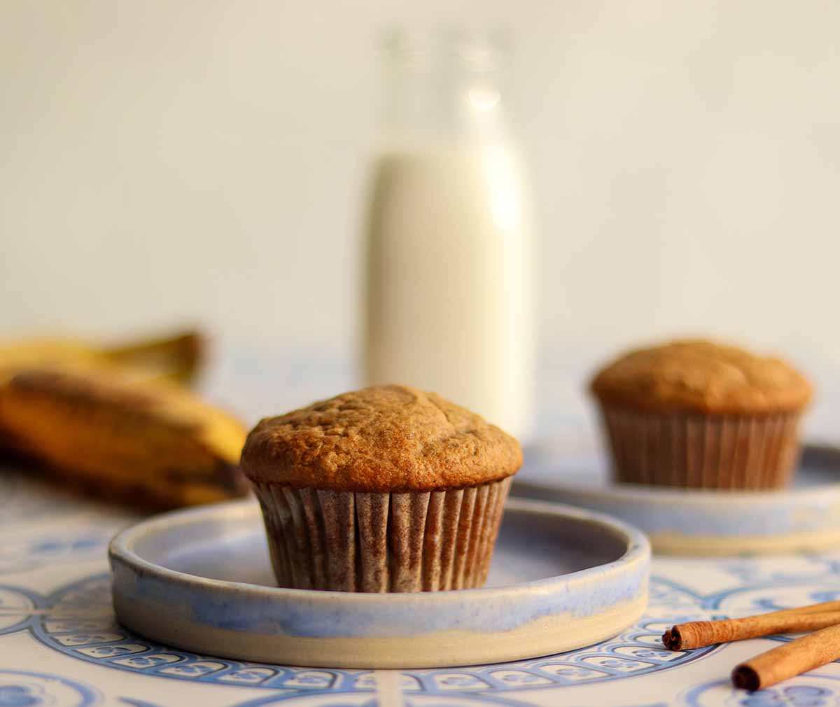 Centered close up of paleo banana muffin on blue ceramic plate surrounded by ingredients and other muffin in background