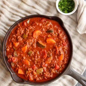 Paleo chili in a cast iron skillet next to a bowl of chives and placed on top of a linen cloth