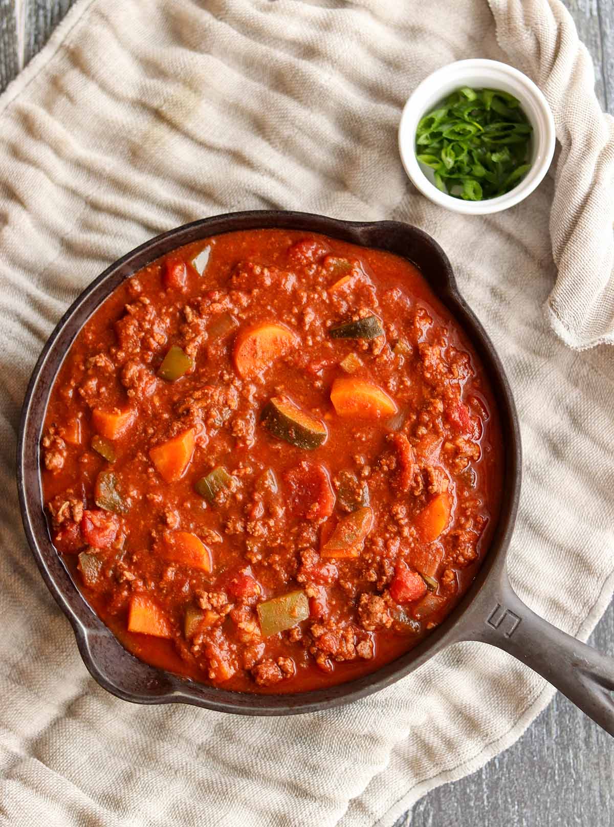 Paleo chili in a cast iron skillet next to a bowl of chives and placed on top of a linen cloth