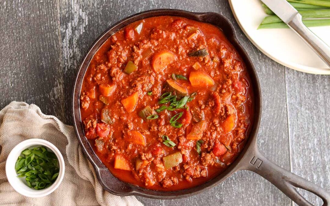 Paleo chili in a cast iron skillet next to some dish wear and cutlery on one side and a bowl of green onions on top of a linen cloth of the opposite