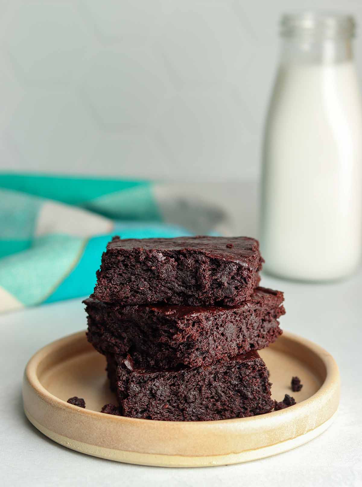 Avocado brownies stacked on top of each other with a blue checkered table cloth in the back ground next to a glass jar of dairy free milk