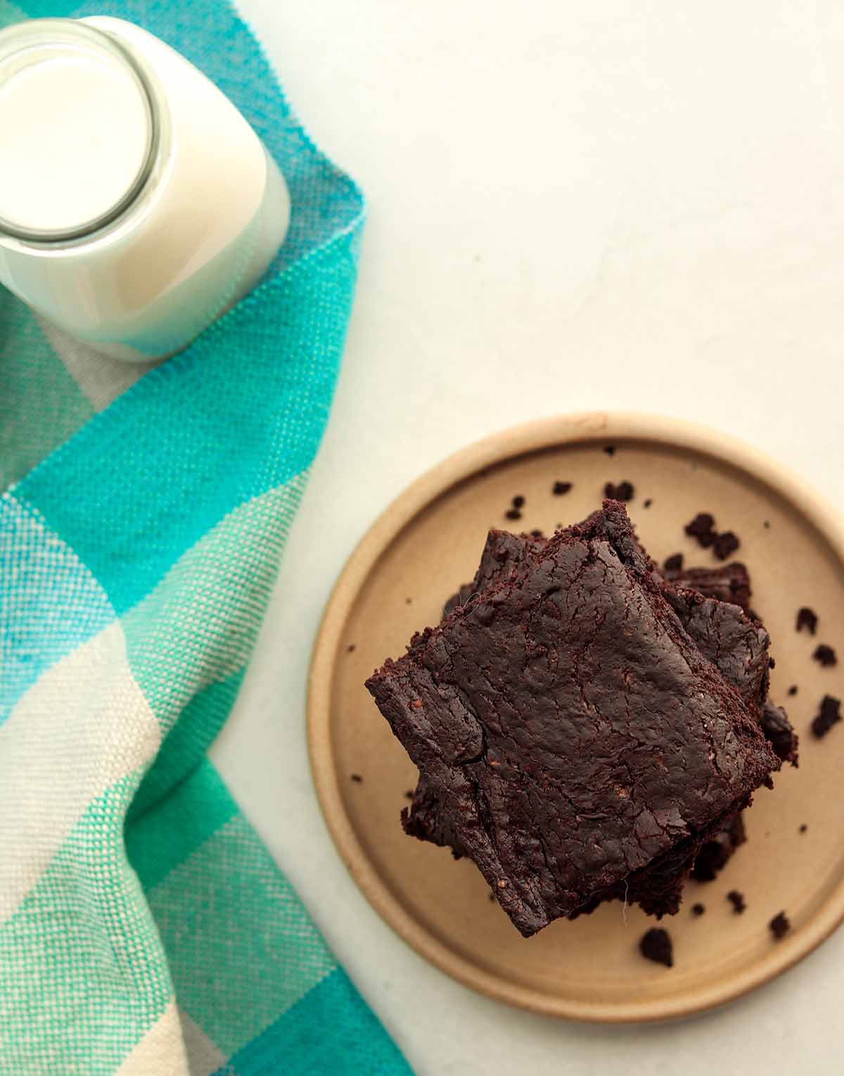 Overhead view of stacked avocado brownies next to a blue checkered table cloth and a glass jar full of dairy free milk