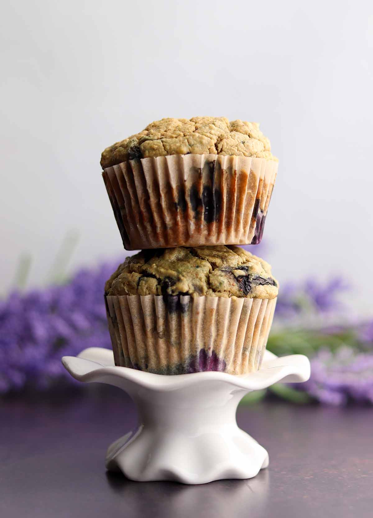 Two blueberry muffins stacked on top of each other on a cup cake pedestal with two bunches of lavender in background 
