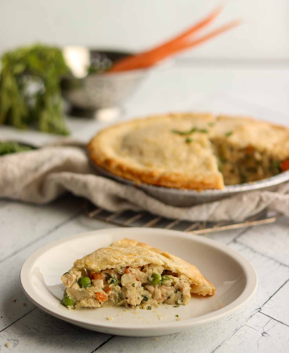 Gluten free dairy free chicken pot pie slice on a place with whole pie in background. Surrounded by fresh ingredients and linen cloth. 
