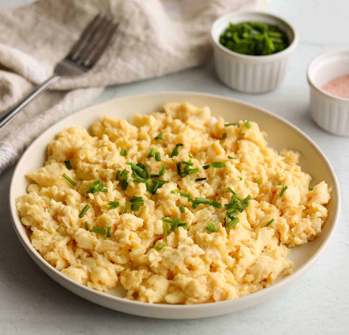 Zoomed out angle of dairy free scrambled eggs topped with green garnish surrounded by a canvas table clothe and fork ready to be devoured. 