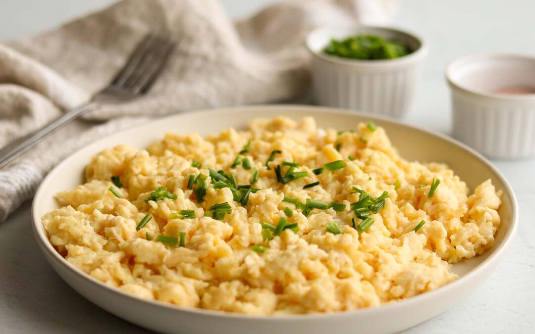 Close of view of fluffy dairy free scrambled eggs topped with green garnish surrounded by neutral coloured kitchen props