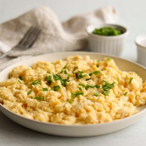 Close of view of fluffy dairy free scrambled eggs topped with green garnish surrounded by neutral coloured kitchen props