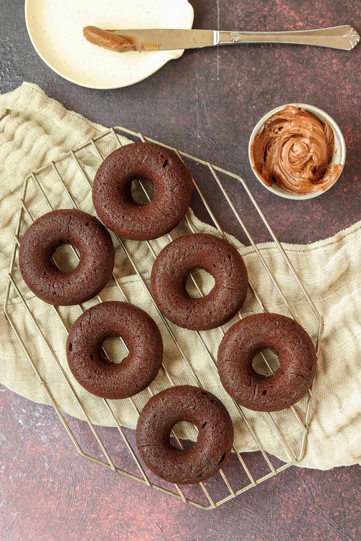 Zoomed out overhead view of chocolate cassava flour donuts on wire cooling rack, placed beside a small dish of toppings